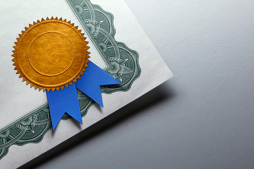 SuperSeva EHS Certification A close up of a gold seal adorned with a blue ribbon is attached to the corner of a certificate of achievement which rests on top of a gray background which provides ample room for copy or text.